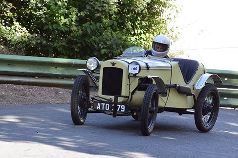 A pale yellow doorless sports two-seater driven fast on a hill-climb road by a helmeted driver. Opens a larger view
