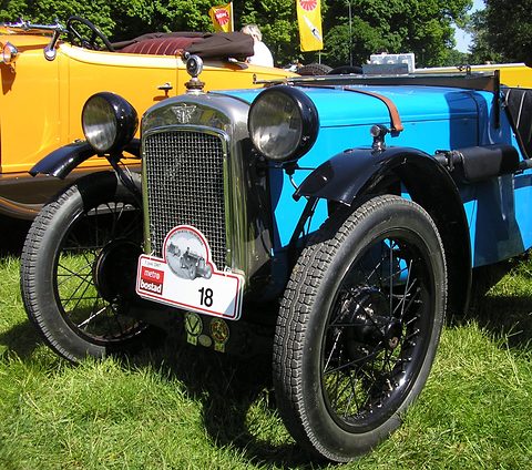 Front quarter of a light blue doorless sports two-seater with a black radiator and rally plate on grass. Opens a larger view
