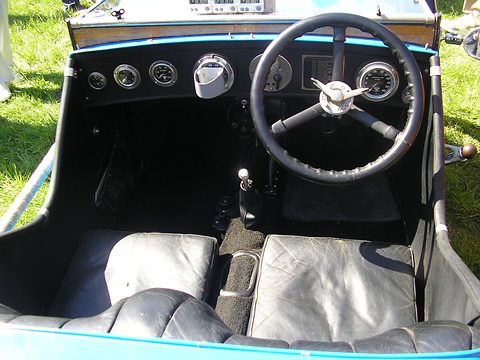 Looking into a narrow cockpit at a black steering wheel, round dials and grey leather seats. Opens a larger view