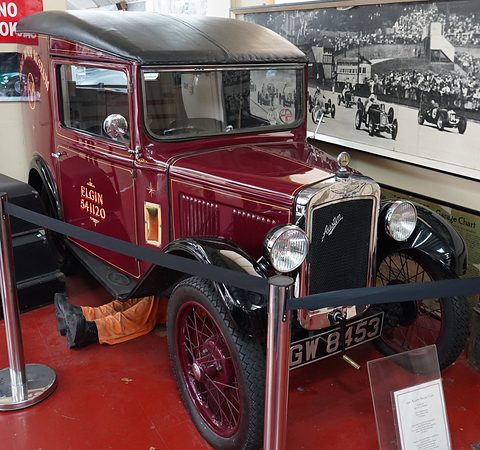 A dark red van with gold lettering and red wire wheels behind a rope barrier in a museum. Opens a larger view
