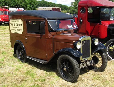 A brown van with trade lettering on its side, black wings and wire wheels on grass beside a red lorry. Opens a larger view