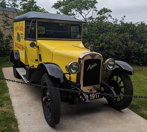 A yellow van with black roof and wings and lettering on its side, parked outdoors behind a chain. Opens a larger view