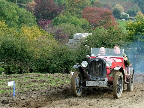A red open special with two crew climbs a muddy slope, exhaust smoke behind it and autumn trees beyond. Opens a larger view