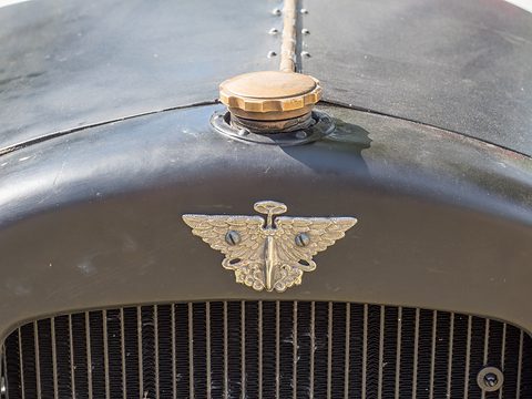 A brass winged badge on a dark green cowl above a radiator grille, a filler cap above it. Opens a larger view