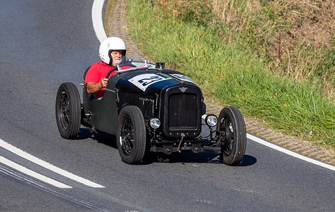 A dark single-seat style racer with a helmeted driver cornering on a hill-climb road by grass. Opens a larger view