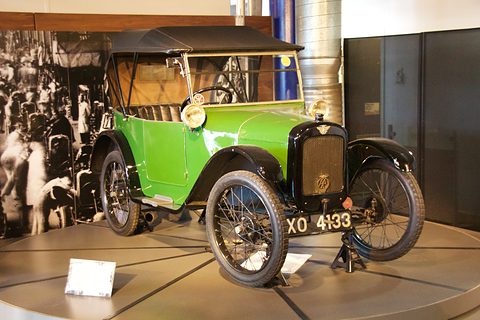 A green open tourer with its hood up, black wings and wire wheels on a turntable before a photo mural. Opens a larger view