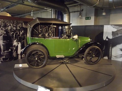 Side view of a green open tourer with its hood up on a round display platform, period photographs behind. Opens a larger view