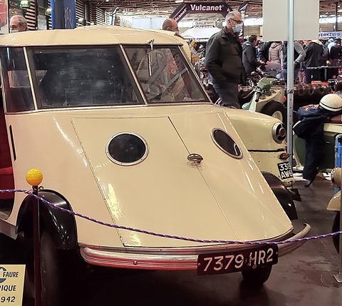 Cream streamlined two-seater with porthole lamps and a split windscreen behind a rope barrier.