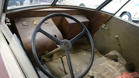 Worn interior with a thin steering wheel, plain wooden boxes and floorboards under a glass screen. Opens a larger view