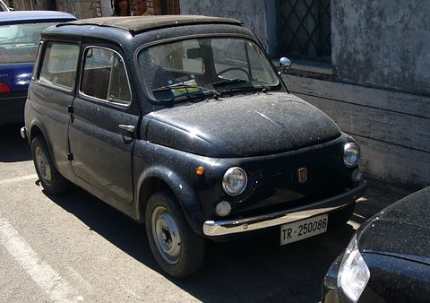 Dusty dark blue small estate car parked in sunshine beside a grey wall with a barred window. Opens a larger view