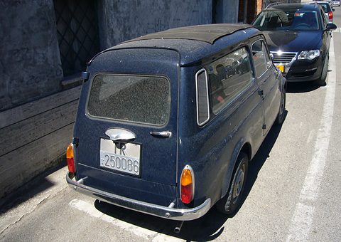 Rear of a dusty dark blue small estate car parked by a wall, a modern saloon behind. Opens a larger view