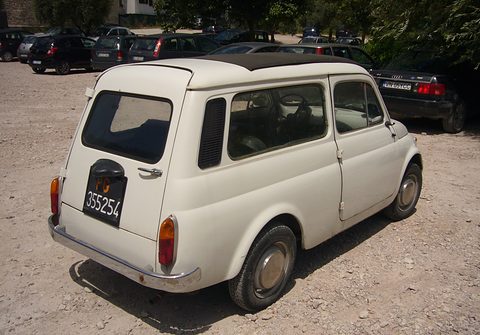 Rear of a weathered white small estate car on gravel, parked cars behind. Opens a larger view