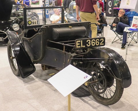Rear of a black three-wheeled vehicle with a leather seat, single rear wheel and number plate. Opens a larger view