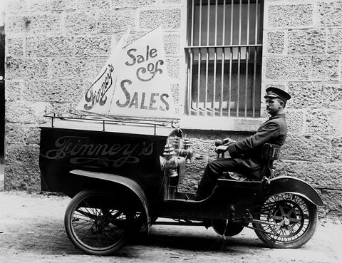 A man in a peaked cap sits behind a box lettered ‘Finney’s’ on a three-wheeled delivery vehicle.