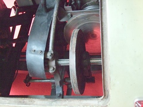 A metal drive disc pressed against a larger flywheel, with a chain guard and shaft, seen through a cut-out in a cream panel.