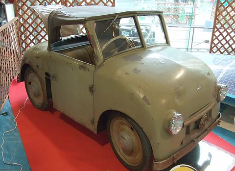 Olive-green open two-seater with a canvas hood, chipped paint and round headlamps, standing on red carpet.