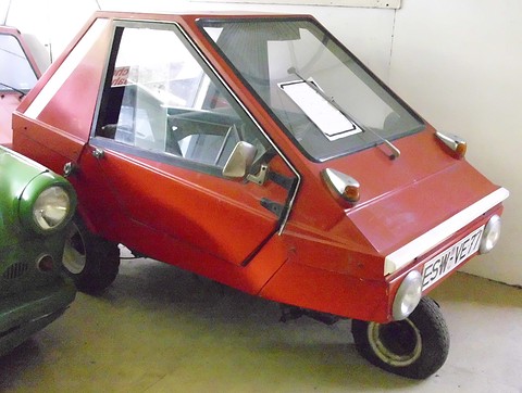Red wedge-shaped plastic microcar with a single front wheel and large side windows, the nose of a green car beside it.