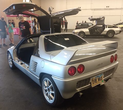 Rear of a silver small coupé with round tail lamps and both doors raised, a DeLorean and people behind. Opens a larger view