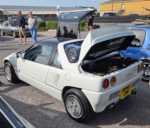 Rear of a white small coupé with its engine cover lifted and a gullwing door raised, people standing behind. Opens a larger view