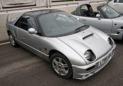 Silver mid-engined gullwing coupé with exposed round headlamps and a bonnet scoop, a UK plate, on tarmac. Opens a larger view
