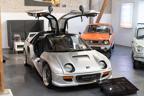 Silver small coupé with both gullwing doors raised beside an information plinth, an orange Mazda behind. Opens a larger view