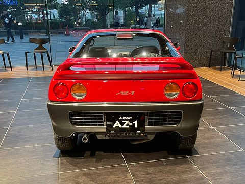 Rear of a red small coupé with round tail lamps, a centre exhaust and an AZ-1 plate on a tiled floor. Opens a larger view