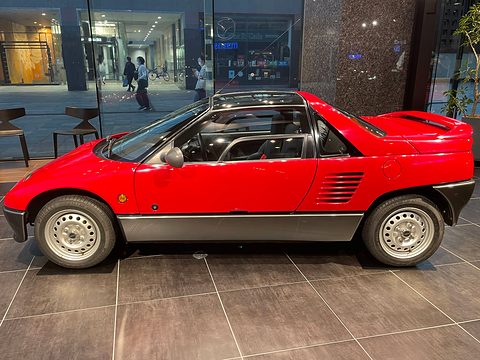 Side view of a red and grey small coupé with slatted side intakes and steel wheels on a tiled floor. Opens a larger view