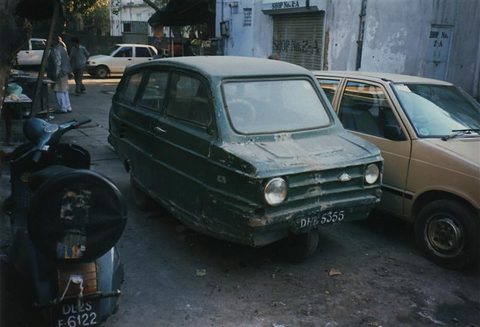 Dark green small three-wheeled saloon with round headlamps parked beside a beige car, scooters in front and shuttered shops behind.