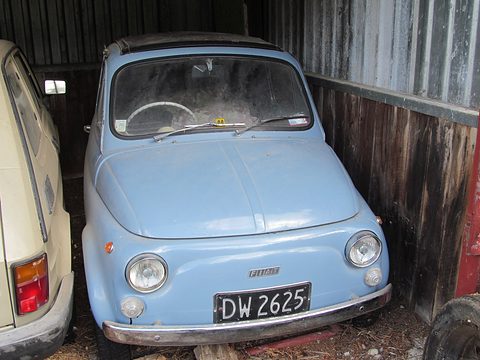 Dusty pale blue Fiat 500 parked in a timber shed, its windscreen grimy, a cream car beside it. Opens a larger view