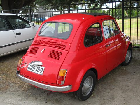Red Fiat 500 from the rear left with a louvred engine lid and New Zealand plate, beside a white saloon on gravel. Opens a larger view