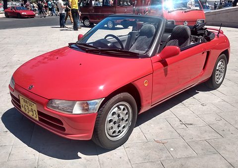 Red small open two-seater with zebra-pattern seats and a Japanese plate on stone paving, people behind. Opens a larger view
