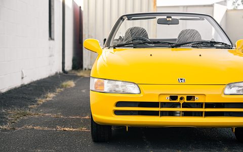 Front of a yellow small open two-seater with slim headlamps parked in a narrow alley between buildings. Opens a larger view