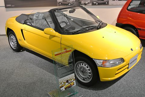 Yellow small open two-seater with its roof folded on a grey floor beside a bilingual information panel. Opens a larger view