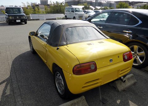 Rear of a yellow small roadster with a black soft top and round tail lamps in a sunny car park. Opens a larger view