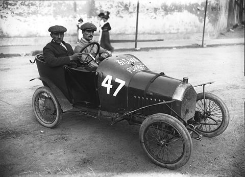 Two men in caps sit side by side in a narrow dark racing car numbered 47 by a wall.
