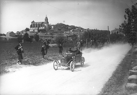 A small open car raises dust on a pale country road below a hilltop village with a church, spectators on the verge.