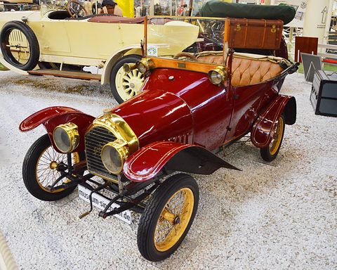Red two-seater with large brass headlamps, brass radiator and yellow wheels on white gravel among other veteran cars. Opens a larger view