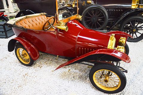 Glossy red two-seater with brass lamps, tan leather seats and yellow-spoked wheels on white gravel, seen from the right. Opens a larger view