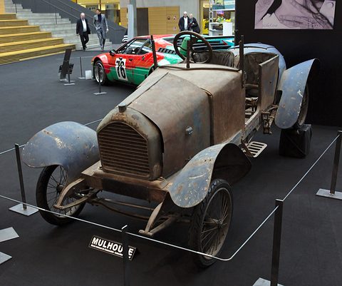 Rusty brown little two-seater with dented blue-grey wings on a black stand, a Mulhouse sign at its foot. Opens a larger view