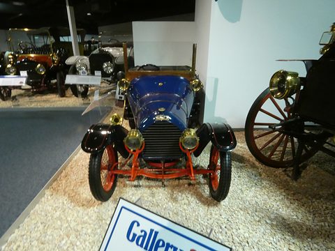 Front of a dark blue two-seater with red-spoked wheels on gravel, a blue sign in the foreground. Opens a larger view