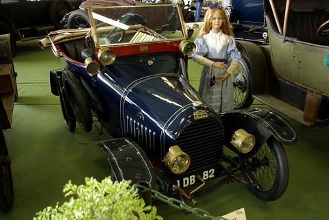 Dark blue two-seater with brass lamps and radiator, a mannequin in a pale period dress standing beside it. Opens a larger view