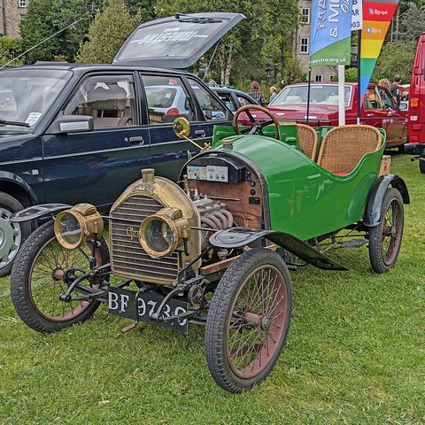 Green two-seater with wicker seats and the bonnet removed showing the engine, on grass with modern cars behind. Opens a larger view