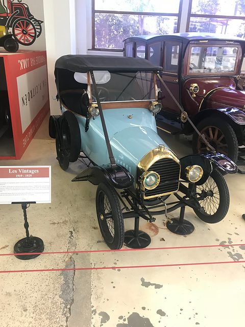 Pale blue two-seater with a black hood and brass radiator on a worn floor, other veteran cars behind. Opens a larger view