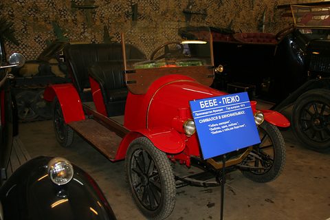 Red open two-seater on a dim studio floor, a blue sign in Russian standing on its bonnet side. Opens a larger view