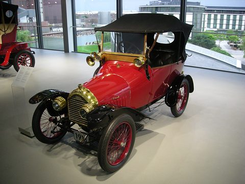 Red two-seater with black hood, brass radiator and red wire wheels on a pale floor by tall windows. Opens a larger view