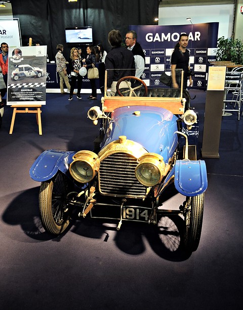 Front of a blue two-seater with big brass lamps and radiator on a dark carpeted stand. Opens a larger view
