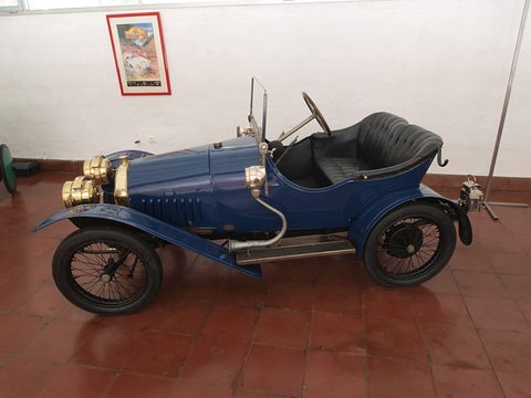 Side view of a dark blue two-seater with a pointed tail, black seats and brass lamps on red tiles. Opens a larger view