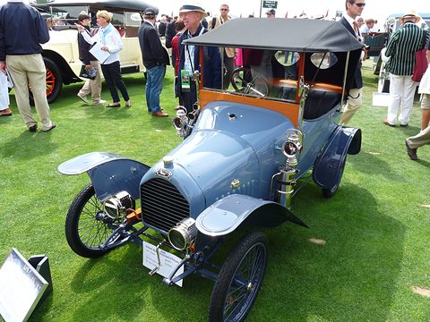 Pale blue two-seater with a black hood and brass lamps on a lawn, spectators walking behind. Opens a larger view