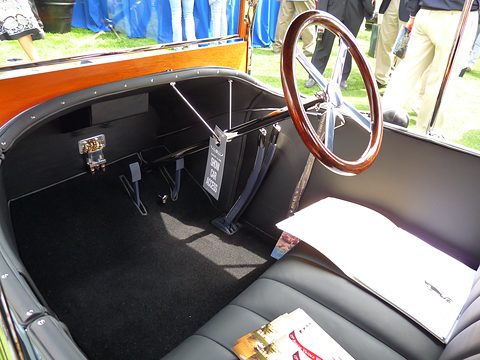 Black leather bench, wooden-rimmed steering wheel, pedals and levers inside a small car, papers resting on the seat. Opens a larger view