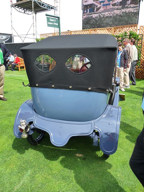 Rear of a small pale blue car with a rounded tail and a black hood with two oval windows, on grass. Opens a larger view